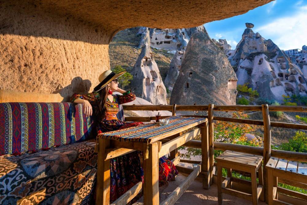 Woman in traditional dress sits on a patterned bench, looking out at rocky, cave-riddled landscape from a carved-out alcove.
