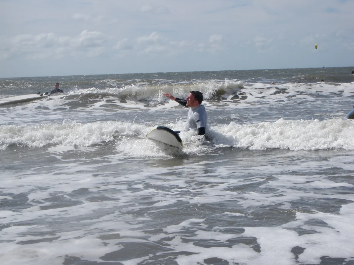 A surfer in a wetsuit rides a small wave, with choppy sea and cloudy sky in the background.