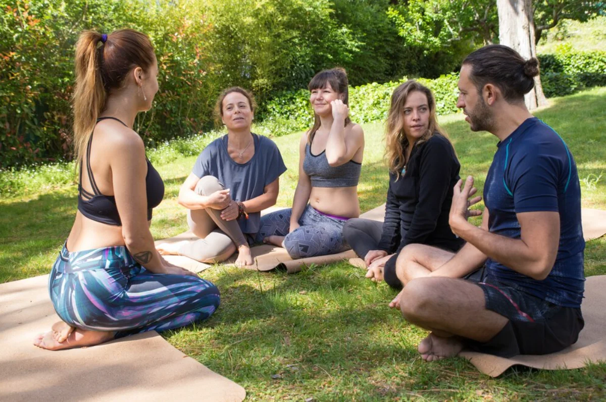 Group of five adults engaged in a casual conversation while sitting on mats in a sunny park.