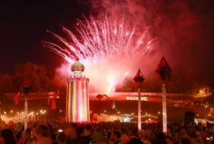 Crowded festival with vibrant fireworks over a colorful tower at night, Glastonbury sign visible.