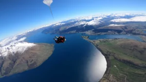 Two people skydiving above a stunning mountainous landscape with snow-capped peaks and deep blue lakes.