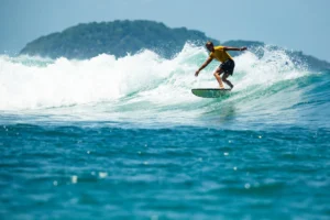 Surfer riding a wave near a hilly island in clear blue waters.