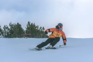 Skier in orange jacket carving through snow, wearing a helmet and goggles, with pine trees and cloudy sky in background.
