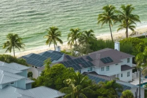 Aerial view of coastal homes with solar panels, near a sandy beach lined with palm trees.