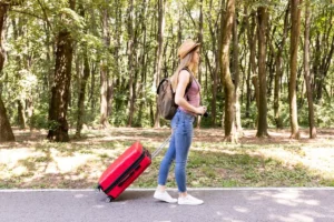 A woman in a hat and backpack walking with a red suitcase on a forest path.
