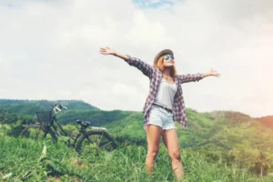 Young woman with arms outstretched by a bicycle in a grassy field, mountains in the background.