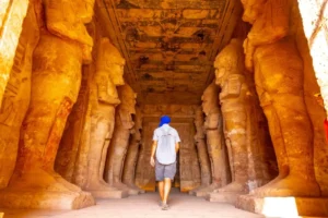 A man in a blue hat exploring a sunlit ancient Egyptian temple lined with large statues.