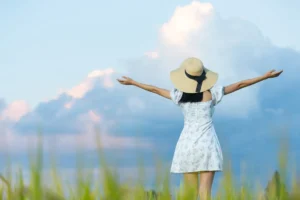 Woman in a summer dress and straw hat, arms spread wide in a lush field under a blue sky with clouds.