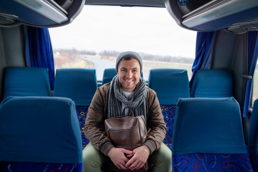Man smiling aboard a bus, seated between blue seats, holding a leather bag, wearing a winter hat and scarf.
