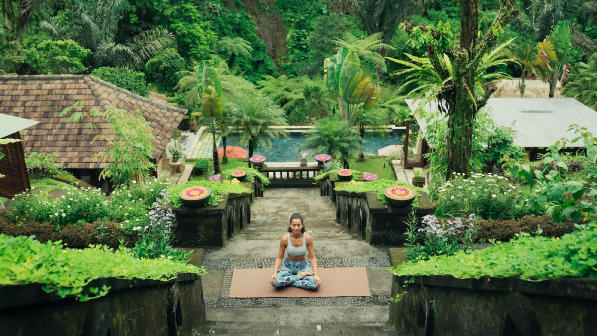 Woman meditating on a yoga mat in a lush, tropical garden with a pool in the background.