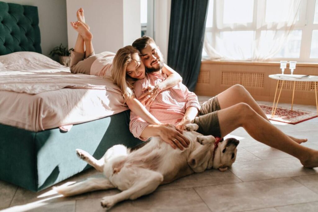 A couple relaxes in a sunny room with their dog, who lies on its back, enjoying their affection.