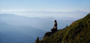Woman meditating on a mountain ledge overlooking a scenic, mist-covered mountain range.