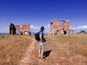 A man with a backpack walking towards ancient ruins in a grassy field under a blue sky.