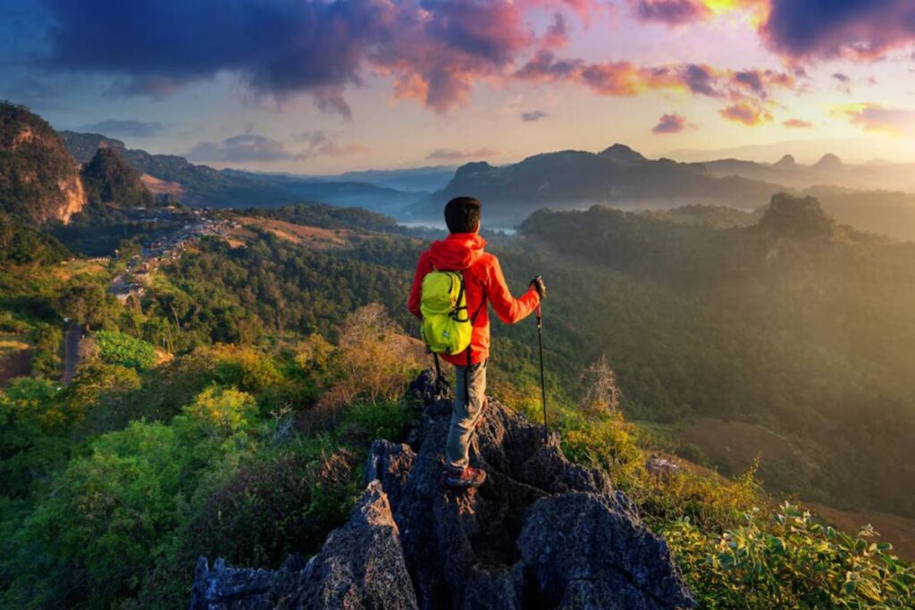 Hiker in red jacket standing on mountain peak at sunrise, overlooking a lush valley.