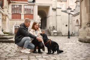 A couple interacts with a black dog on a cobblestone street in a quaint old town.