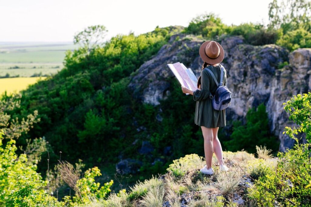 Woman in hat and backpack reading a map on a rocky hillside with lush greenery.