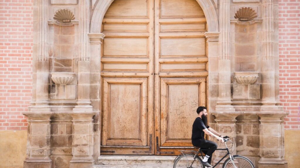 A man cycling past an old, grand wooden door with stone architectural details.