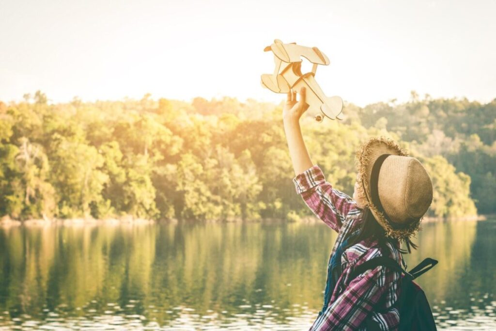 Person in hat holding a toy airplane aloft by a lakeside, surrounded by forest, in warm sunlight.