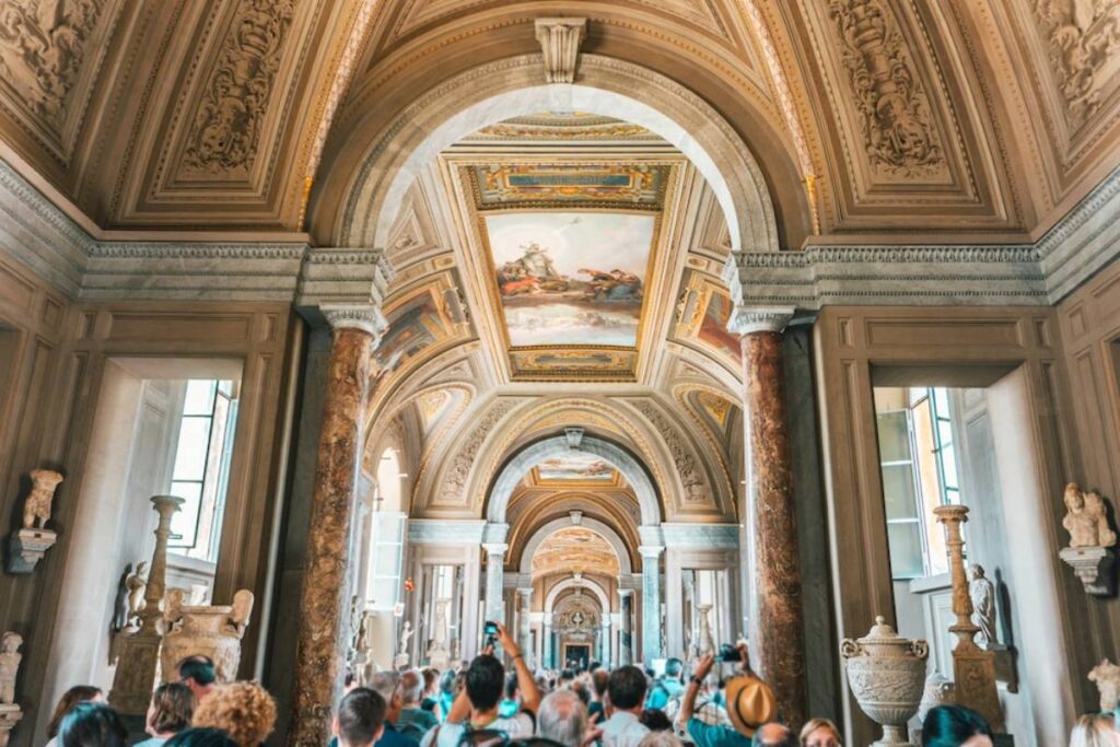 Crowd of tourists inside an ornate museum hallway with classical sculptures and intricate ceiling frescoes.